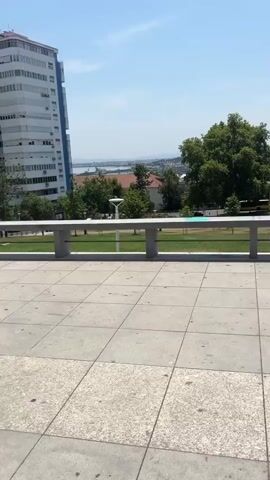 The video pans across a public space. In the foreground, there is a tiled patio with a concrete railing. Beyond the railing, there is a grassy park with trees and some park benches. In the distance, there is a large, modern building with blue accents and further out, a body of water with boats and a city skyline. The sky is clear and blue. There are no people clearly visible in the park area but one woman is seen walking up stairs at the end of the video. The overall impression is of a bright, sunny day in an urban park setting.