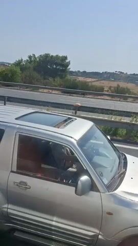 The video shows a view from inside a vehicle traveling on a highway. The camera pans to the right, revealing a silver SUV in the adjacent lane. Further ahead, on the same highway, a small yellow and white paddle boat is being transported on a trailer behind a pickup truck. The paddle boat appears to be old and weathered. Other vehicles, including a blue semi-trailer truck and several cars, are also visible on the highway. The landscape outside the highway consists of rolling hills with sparse vegetation under a clear blue sky. The footage is shaky, indicating movement of the vehicle.