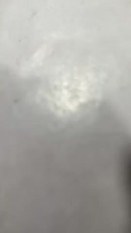The video shows a close-up of a green fidget spinner with a red circle and a white star in the center, placed on a white surface. The spinner is spinning. The background is out of focus and appears to be a desk or table with papers and other objects. The lighting is bright, creating reflections on the spinner.