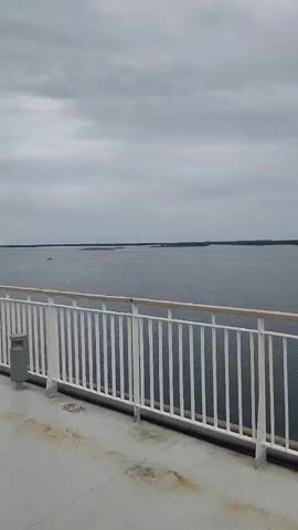 A video shot from a ship deck shows a vast expanse of water under a cloudy sky. A white railing with wooden accents runs across the foreground, separating the deck from the water. A gray trash bin is visible to the left of the frame, and a speaker is mounted on a pole to the right. In the distance, a long, thin landmass stretches across the horizon. A faint, bright horizontal line, possibly a reflection or a light source, is visible in the upper right quadrant of the sky. The water appears calm and dark, and the overall atmosphere is subdued due to the overcast weather. No people are visible in the footage.