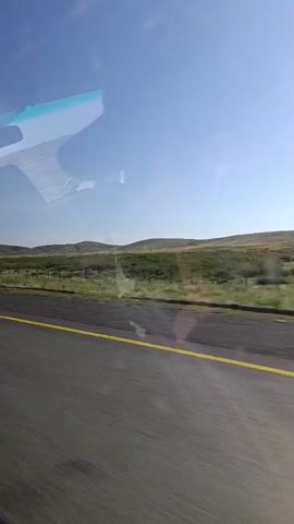 The video shows a view from inside a vehicle traveling on a paved road with a yellow line. The road is in the foreground and is blurred, indicating motion. Beyond the road is a fence, then a grassy field with rolling hills in the background. The sky is clear and blue. There is a reflection of the interior of the vehicle on the windshield, showing parts of the dashboard and a person's head. A large blue truck passes by on the right side of the frame, its wheels and side visible. The video is taken from a moving vehicle.