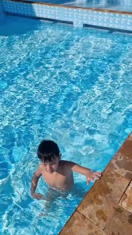 A young boy is in a swimming pool. He is partially submerged in the water and is holding onto the edge of the pool with his right hand. He is looking towards the camera. The water is clear and blue, and the sunlight is reflecting off the surface. The boy is wearing swim trunks. The pool has a tiled edge.