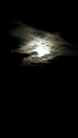 The video shows a nighttime sky with a bright moon partially obscured by dark clouds. The moon's light illuminates the edges of the clouds, creating a dramatic effect against the blackness of the sky. The scene is still and silent, with no visible objects or people.