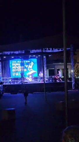 A nighttime outdoor stage setup is visible. A large blue screen displays text and an image of two people dancing. A drum set is positioned in front of the screen. Three individuals are on the stage, one near the drum set and two further to the right. In the foreground, a person walks across the dark ground. Several cube-shaped objects are scattered on the ground. The scene is lit by stage lights, creating a predominantly blue hue.