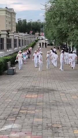 A group of people are walking and exercising outdoors on a paved walkway lined with trees. The people are mostly dressed in white clothing, appearing to be in a coordinated group activity. Some individuals are walking at a moderate pace, while others seem to be engaged in more dynamic movements. The environment suggests a park or an outdoor recreational area, with buildings and a street visible in the background.