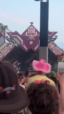 The video shows a crowd of people at what appears to be an outdoor festival or event. In the background, there is a large stage structure with a distinctive design, possibly featuring a large face as part of the decoration. Several balloons are floating in the air, including some shaped like animals such as a cat and a bee, and some round balloons in various colors. People in the foreground are mostly facing away from the camera, towards the stage. Some individuals are wearing hats, and one person has pink cat ears on their head. The lighting suggests it is daytime. The overall atmosphere seems energetic and festive.