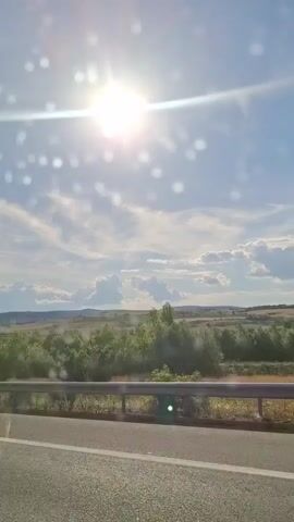 This video captures a view from a moving vehicle. The sun is bright and positioned in the upper left quadrant of the sky, casting lens flares and creating bright spots throughout the frame. The sky is blue with scattered white clouds. The foreground consists of a paved road with a white line marking the lane. A metal guardrail runs parallel to the road, separating it from the landscape beyond. The landscape features rolling hills covered in green vegetation and trees. The movement of the vehicle causes a slight blur and suggests a journey through a natural environment under a clear, sunny sky.