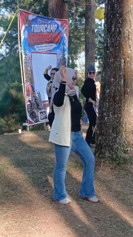 A group of three women are dancing outdoors. The woman in the foreground, wearing a headscarf, black shirt, white vest, and blue flared jeans, is center frame and facing the camera. She is smiling and has her hands in a dancing pose. Behind her and to the left, another woman in a black shirt and jeans, also wearing a headscarf, is dancing and has her right arm raised. To the right of the center woman, a third woman wearing sunglasses and a black t-shirt with a graphic design, is partially obscured by a tree and appears to be dancing with her right arm extended. A banner with the text "TOURCAMP KEMERDEKAAN" is visible behind the woman in the foreground. The environment is grassy with trees and dappled sunlight.