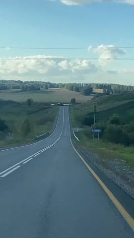 A first-person perspective video shows a vehicle traveling on a paved road through a rural landscape. The road has a double white line marking the center and a yellow line along the right edge. The road curves slightly to the left and then straightens, heading uphill towards the horizon. Rolling green hills with scattered trees and fields stretch out on either side of the road. In the distance, a line of trees forms a dark silhouette against a bright blue sky dotted with white, fluffy clouds. A few utility poles are visible along the right side of the road. A blue sign with white text is mounted on a pole on the right side of the road. The lighting suggests it is daytime, likely late afternoon or early evening due to the warm tones in the sky. There are no people or other vehicles visible in the frame.