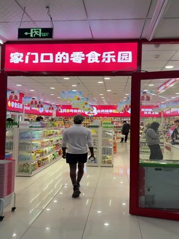 The camera pans from the entrance of a well-lit grocery store. The entrance is marked by a red sign with white text in Chinese, and a green exit sign. A Black man wearing a light-colored long-sleeved shirt, black shorts, a dark baseball cap, and sneakers walks into the store. Inside, rows of shelves are stocked with various food items and beverages. Several other customers are browsing the aisles. The store has a clean, modern appearance with glossy white tiled floors. Promotional banners and posters hang from the ceiling. A checkout counter is visible towards the back of the store with a female customer standing near it.  The video ends with the camera focusing on the checkout area.