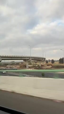 A view from a moving vehicle shows an overpass and cars on a highway. The sky is overcast. The motion blur indicates speed. There are trees and construction equipment visible in the distance. The video pans to show more trees and foliage.
