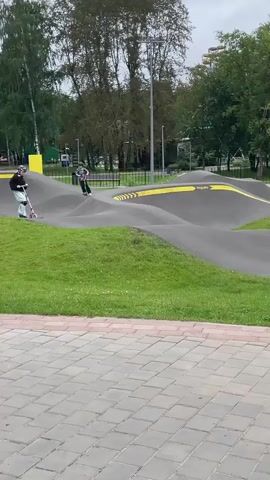 A group of children wearing helmets are riding scooters on a pump track. The track is made of asphalt with yellow markings and is surrounded by grass. In the background, there are trees and some buildings. The weather appears to be overcast.