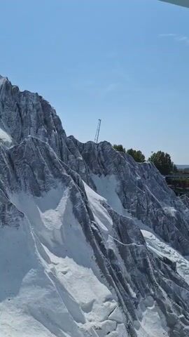 A handheld vertical video pans across a snowy mountain landscape. In the foreground, a sharp, gray, rocky mountain face is covered in patches of white snow, giving it a jagged, textured appearance. A thin, metallic pole or antenna is visible jutting out from the summit of the mountain. Beyond the mountain, a sprawling miniature village or park unfolds under a bright blue sky with scattered clouds. Numerous tiny buildings, greenery, pathways, and a body of water are discernible. Small figures of people are dotted throughout the park, suggesting it is a public attraction. The video is captured from an elevated perspective, possibly from a nearby structure or attraction.