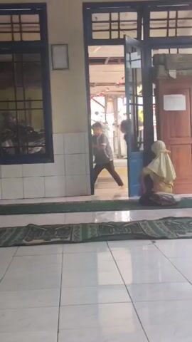 A young boy exits a doorway and walks towards the camera. He is wearing a gray hoodie and dark pants. He walks past a woman who is sitting on the floor facing away from the camera. The woman is wearing a light-colored hijab and a yellow top with dark pants. She is sitting on a prayer mat. The floor is tiled white and there are prayer mats laid out. The doorway leads into a room with a wooden ceiling. There are windows with blue frames on the wall to the left of the doorway, and another window on the wall to the right. The exterior of the building is visible through the windows, which appears to be an outdoor area.