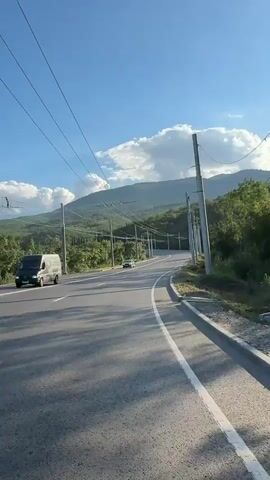 The video shows a car driving on a winding road that goes uphill towards mountains. The sun is shining brightly, and there are some clouds in the sky. There are also telephone poles and wires along the side of the road, and trees and greenery on the hills. The road has white lines marking the lanes. The car is gray and is driving in the left lane. The camera appears to be mounted on a vehicle or held by someone in the passenger seat of a vehicle.