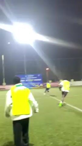 The video shows a group of people playing soccer on a green artificial turf field under bright artificial lights at night. Some players wear yellow vests, while others wear red shirts. The camera follows a person in a yellow vest and black pants as they walk along the sideline. There are banners and a fence visible in the background, with some buildings and trees behind the fence. The players are actively running and kicking the ball, passing to one another.