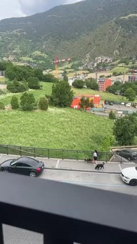 A high-angle daytime video captures a woman walking two dogs on a leash along a sidewalk in Encamp, Andorra. The woman, with long dark hair, wears a light top and dark shorts. One dog is medium-sized, fluffy, and multi-colored (grey/brown), while the other is a black Labrador-type dog. They walk past a black two-door car, possibly an Audi TT, parked parallel to the sidewalk. A white car is partially visible parked further down on the right.

Beyond a black metal fence, a large, vibrant green grassy field stretches out. In the mid-ground, several buildings with orange roofs are visible, along with orange construction containers and a yellow crane, indicating ongoing development. The background is dominated by lush green mountains under an overcast sky, with more buildings scattered across the valley. The road in the foreground has blue markings.