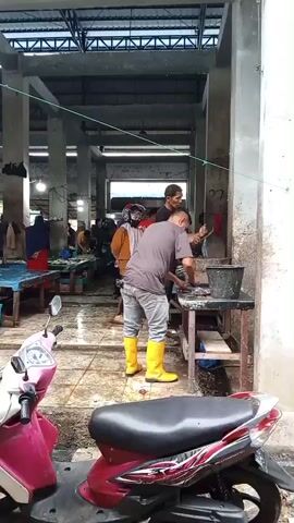 The video shows a bustling market scene, likely a fish market, given the presence of what appear to be fish being handled. In the foreground, a red and silver scooter is parked. The main activity is centered around a table where two men are actively engaged in preparing fish. One man, wearing a grey t-shirt and light-colored pants, is wearing bright yellow boots and is leaning over the table, seemingly washing or cleaning fish. Another man, in a black t-shirt, stands beside him, also appearing to be involved in the task.

In the background, other market-goers can be seen. A woman in an orange top and headscarf is visible, as are other individuals moving around the stalls. The setting is an open-air structure with a metal roof supported by concrete pillars. The ground appears to be wet, suggesting either recent rain or the ongoing cleaning of the market. The overall atmosphere seems busy and functional, typical of a working market. There is no clear indication of the time of day or weather conditions beyond the wet ground. A number "27" is visible on a pillar in the background. The location is identified as Puloara, Indonesia.