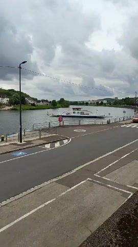 Here is a description of the video. 
The video shows a view of a river from an elevated perspective, with a ferryboat docked along the bank. The river is surrounded by buildings and trees on both sides. The sky is overcast with dark, heavy clouds. The road in the foreground is paved, with white lines marking the lanes. There is a designated parking space for people with disabilities on the left. A lamppost stands next to the road. The overall tone of the video is calm and peaceful.