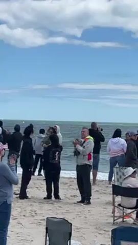 Here is a description of the scene in the video:

The scene takes place on a sandy beach on a partly cloudy day. A crowd of people stands along the shoreline, facing the water. Most are standing, though a couple are sitting in beach chairs. The group is applauding. Some individuals are taking photos with cameras.

The ocean waves are visible in the background, and there appears to be a marker or buoy bobbing in the water.  The sky is a mix of blue and white, with scattered clouds. The attire of the people varies.