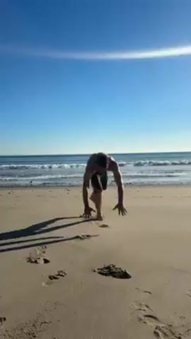 A person is performing a handstand on a sandy beach. They are facing away from the camera, towards the ocean. The person is shirtless and wearing shorts. The sky is blue and clear. The ocean is visible in the background, with small waves. There are footprints in the sand around the person. After holding the handstand for a few seconds, the person slowly comes down, bending their legs and landing on their back.