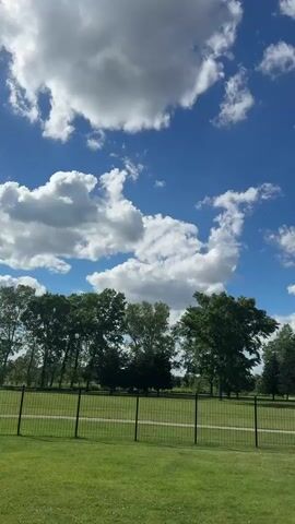 The video captures a bright, sunny day with a vivid blue sky dotted with fluffy white clouds. In the foreground, a well-maintained green lawn stretches out, bordered by a black chain-link fence. Beyond the fence, a line of lush green trees creates a natural backdrop. The camera pans slowly from left to right, offering a panoramic view of the park-like setting. The scene is serene and peaceful, with no people or animals visible. The overall impression is one of natural beauty and tranquility.