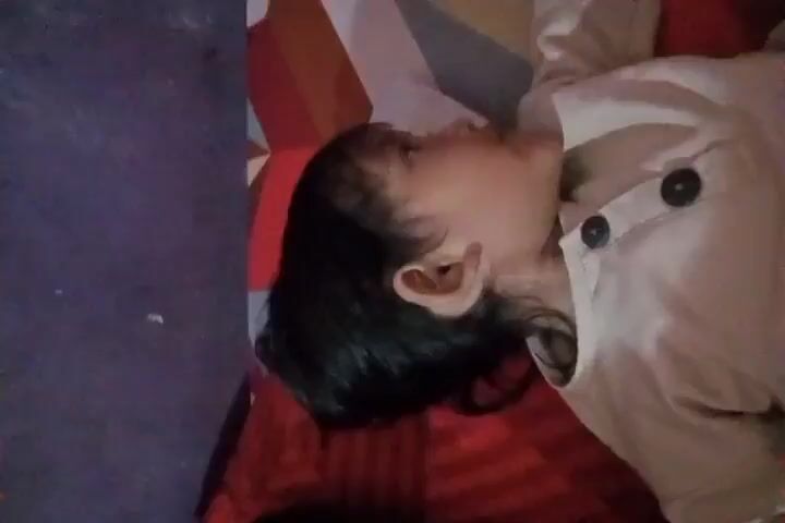 A close-up video shows a young child lying on their back, looking at the camera. The child is wearing a light-colored shirt with dark buttons. Their eyes are open and they appear to be talking or singing. The background shows a colorful, patterned surface and a red pillow. The video is shot vertically and is slightly shaky.
