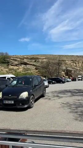 The video shows a row of parked cars on an asphalt lot, with a rocky cliff face rising in the background. The sky is blue with scattered clouds. The cars include a black hatchback in the foreground, a silver Mercedes-Benz with the license plate TT-653-TN, and several dark-colored vans. In the distance, a group of people are gathered near a white van. A leafless tree stands between the cars and the cliff.