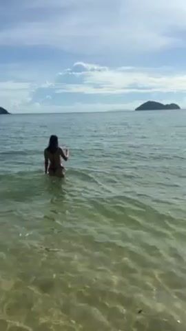 A woman in a dark bikini walks into the ocean. The water is shallow and clear near the shore. An island is visible in the distance. The water reflects the sunlight, creating a shimmering effect. A jet ski appears in the distance.