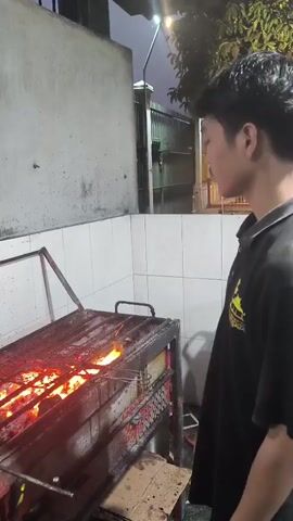 A young man wearing a black polo shirt is standing in front of a grill. The grill is large and appears to be made of metal. There are flames coming from the grill, indicating that food is being cooked on it. The man is holding a metal skewer with food on it over the flames. He is looking at the grill. The background of the scene is a wall that is partially covered in white tiles and partially made of concrete. There is also a fence visible in the background. The lighting suggests it is evening or night.