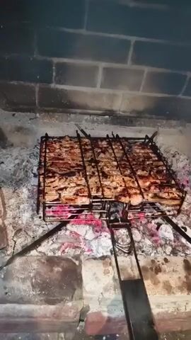 The video shows a grill basket filled with meat being cooked over hot coals in a fireplace. The meat is browning and appears to be cooking evenly. The fireplace is made of brick and stone, and there is smoke rising from the fire. The camera angle is high and looking down at the grill.