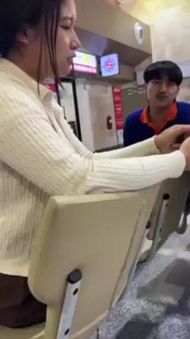 A woman holds a smartphone showing a video on the TikTok app, in front of two employees wearing blue and orange uniforms. The video displays a first-person view of a small, open-air train traveling on tracks through greenery. The woman's hand covers the bottom of the phone, obscuring some of the screen. The employees seem to be watching the video with interest. The setting appears to be an indoor waiting area, possibly an airport, with tiled flooring and a check-in counter visible in the background.