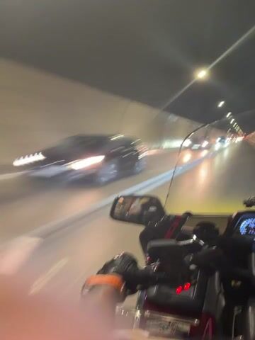 A first-person perspective video shows a motorcycle traveling through a tunnel at night. The tunnel walls are lit with overhead lights and appear to be made of concrete. Cars are also visible in the tunnel, with their headlights and taillights creating streaks of light due to motion blur. The motorcycle has a windshield, rearview mirrors, and controls that the rider's gloved hands are operating. The rider's arm is visible, with tattoos on the forearm. The overall scene is dark and illuminated by artificial lights, creating a sense of speed and movement.