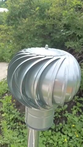 The media file shows a close-up, eye-level view of a metal turbine vent located outdoors. The turbine, which is silver and has a spherical shape with spiraling blades, sits atop a metal pipe. The background consists of lush green foliage, suggesting a natural or garden setting. The lighting indicates it is daytime, and the foliage appears healthy, implying favorable weather conditions, possibly spring or summer. The focus is on the turbine vent itself, highlighting its metallic texture and the way its blades are designed to catch the wind. There is no visible text or people in the media file. The setting, with its dense greenery, suggests an outdoor environment, likely in a residential or rural area given the presence of what appears to be a ventilation component. The overall mood is neutral, focused on the object and its surroundings.