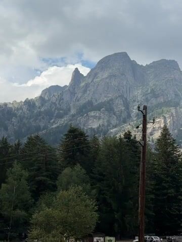 The video shows a landscape view of a mountain range behind a forest and a field with some parked vehicles. In the foreground, a wooden utility pole with electrical wires is visible. The sky is cloudy, and the overall lighting suggests it might be overcast or late afternoon. The mountains are rocky and rugged, with some vegetation on their slopes. The forest consists of various types of trees, mostly evergreen. In the field, there are several RVs and cars parked, indicating a possible campsite or rest stop. The scene appears to be a natural environment with signs of human presence.