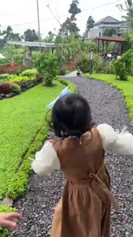 Two young girls are running along a gravel path in a garden.  The girl in front is wearing a brown dress, a white long-sleeved shirt, and a blue bow in her hair.  The girl behind her is wearing a light blue dress with a white, textured top.  The path is lined with green grass and small stones, and there are trees, plants, and a gazebo visible in the background.  The sky is overcast.