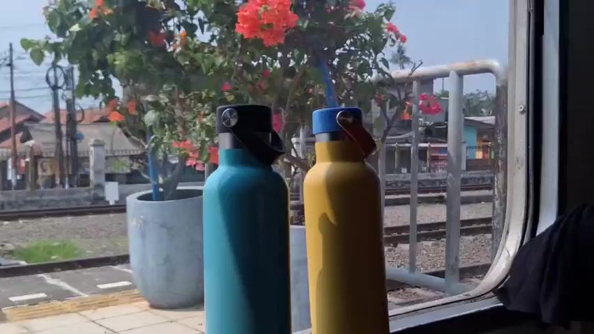 The scene shows a view from inside a moving train through the train window.  Two insulated water bottles, one turquoise and one yellow, stand side-by-side on a surface within the train. Through the window, railroad tracks are visible, along with various buildings of low to medium height. The buildings are a mix of colors and appear to be in a residential or light industrial area. The sky is blue and the overall lighting is bright.