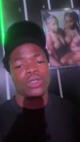A dark-skinned man is video recording himself with a smartphone in a room with changing lighting. The background consists of black and white vertical panels and an framed picture on the wall. The man is wearing a black shirt and a black cap. The lighting in the room shifts from red to green to purple, casting different hues on the man's face. He speaks in a way that looks like he is rapping and also moves his head and hands, dancing to the music playing.