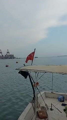 The video shows a white fishing boat docked at a pier. The boat has a covered canopy, fishing nets, and a Turkish flag at the front. In the background, there are large cranes, ships, and a body of water. The sky is overcast. The camera pans across the boat, showing its deck and equipment.