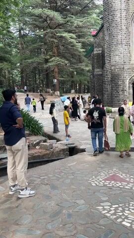 The video shows a wide shot of a stone church with several people standing outside. The church is made of gray stone and has a pointed arch over the doorway. Tall trees are visible around the church. People of varying ages and ethnicities are milling around, some entering the church and others standing in small groups. The ground is paved with stones, and there are some green plants and shrubs visible in the foreground. The sky is overcast.