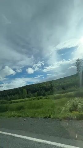 The video shows a scenic view from inside a vehicle, likely a car, looking out at a landscape. The sky is partly cloudy with the sun shining brightly, casting rays onto the scene. The landscape consists of rolling green hills covered in dense trees. In the foreground, there is a road with white lines. A power line tower is visible on the left side of the frame. The vehicle appears to be moving, as suggested by the slight blur of the roadside elements and the perspective shift. At one point, the rear of a silver SUV with a license plate that reads "C221KV 74" is seen in the frame. Further along, the front of a white car is visible. The overall impression is a drive through a natural, possibly rural or mountainous, environment on a sunny day.