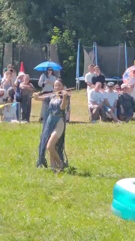 A young woman in a flowing blue dress plays the violin in a grassy field. She is barefoot and her dress has long, thin fringe. She is facing the camera and appears to be performing for a crowd. Behind her, several people are sitting on benches and chairs, some are holding umbrellas. There are trees and foliage in the background. The scene appears to be outdoors in daylight.