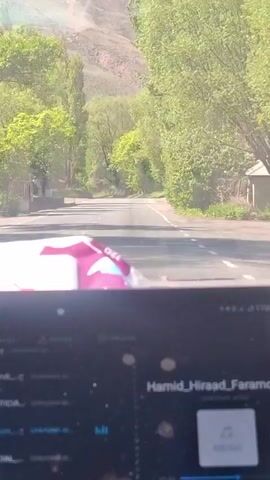 A car drives along a straight, empty road, lined with green trees on either side. A large screen in the foreground displays music playing by Hamid Hiraad Fan. A pack of baby wipes is visible above the screen. In the distance, a mountain can be seen. The day appears sunny.
