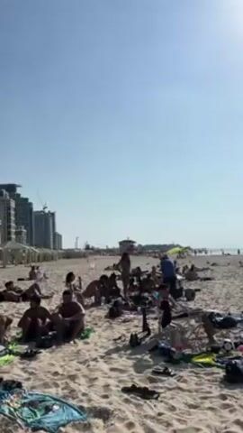 The video shows a crowded beach on a sunny day. People are sitting, lying down, and walking around on the sand. Some are sunbathing, while others appear to be talking or relaxing. In the background, there are tall buildings and a lifeguard tower. The water is visible, with people swimming and enjoying the sea. There are personal belongings scattered around on the sand, such as bags, towels, and shoes. The sky is clear and blue.