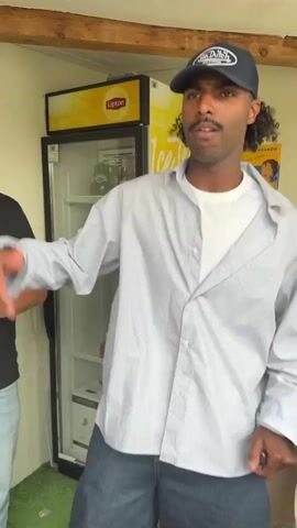 A man wearing a Van Dutch baseball cap, a white t-shirt under an unbuttoned light-blue striped shirt, and wide-legged jeans is dancing. He is standing in front of a cooler with a Lipton Ice Tea advertisement on it and a vintage poster of a woman. The man is smiling and gesturing with his hands as he dances. There are other people in the background partially visible. The scene appears to be outdoors in a shaded area.