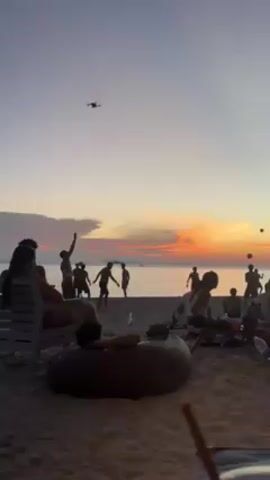 A beach scene at sunset features a group of people playing beach volleyball, while others relax on chairs and a beanbag. A drone hovers in the air, presumably filming the scene. The sky is a mix of orange, pink, and purple hues, suggesting a vibrant sunset. The beach is sandy, and the sea is visible in the background.