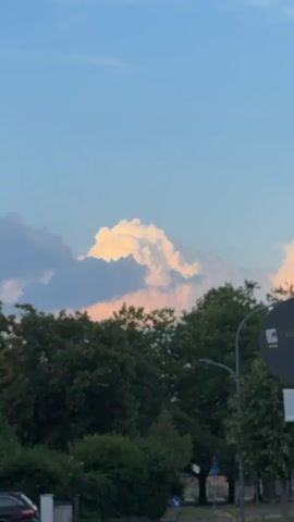 The video shows a close-up view of a cloudy sky at sunset. The clouds are illuminated by the setting sun, creating a dramatic effect of light and shadow. The clouds are a mix of white, yellow, and pink hues, with some darker, purplish clouds in the foreground. The sky above is a clear blue. In the lower part of the frame, the tops of green trees are visible, suggesting an outdoor setting. The camera is focused on the clouds, with the trees serving as a grounding element. The video is a static shot, with no movement other than the subtle changes in light on the clouds.