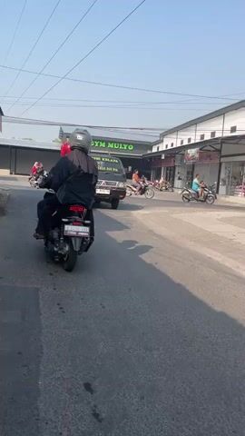 The video begins with a person on a motorcycle, wearing a black jacket and a white helmet, riding on an asphalt road. The motorcycle's brake light is illuminated. To the right of the motorcycle, a black Mitsubishi L300 pickup truck is driving forward. The truck has a white sign on the back with yellow lettering that reads "UD. BUMI KELAPA". There is also a yellow sticker on the windshield. On the truck bed, there are stacked white rectangular objects, possibly bricks or building materials. In the background, on the left side of the road, there is a building with a sign that says "LOSS GYM MULYO" in green lettering. The building appears to be a commercial establishment with glass doors and windows. To the right of the "LOSS GYM MULYO" building, there are several other storefronts. Further down the road on the right, another person is riding a motorcycle. The sky is clear and blue, indicating daytime. The road surface is asphalt with some cracks and wear marks. The overall scene depicts a street in what appears to be an urban or commercial area.