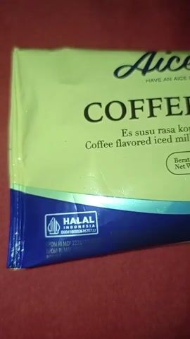 A close-up, slightly blurry shot of the packaging for an Aice brand iced milk bar. The packaging is primarily light green with a blue and purple wave design at the bottom. Prominently displayed are the words "Aice" in script, "COFFEE CRISPY" in large capital letters, and a description in Indonesian and English stating "Es susu rasa kopi lapis cokelat kacang" and "Coffee flavored iced milk coated with peanut chocolate." A "HALAL INDONESIA" certification is visible in the lower left corner. To the right, a circular emblem reads "TOP BRAND 2024". The net weight is indicated as 45g. The background is a red, textured surface, likely fabric. The lighting is direct and appears to be from above.