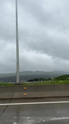 The video shows a highway scene on a cloudy day. A concrete barrier with yellow reflectors lines the road, separating it from a grassy area with wildflowers. In the background, hazy mountains stretch across the horizon under an overcast sky. The road surface is wet. The camera appears to be positioned inside a moving vehicle.